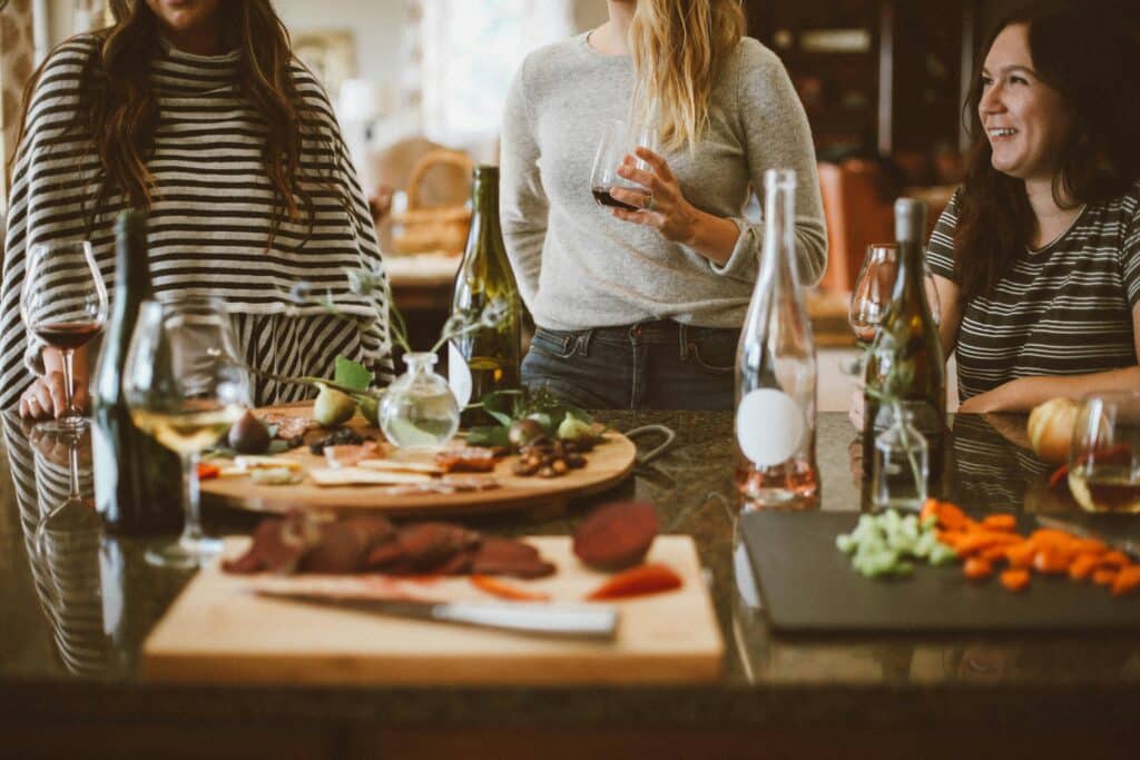 Três mulheres em uma mesa com tábuas de vinho e charcutaria. Duas mulheres estão de pé, com as cabeças cortadas da imagem, e uma está sentada.