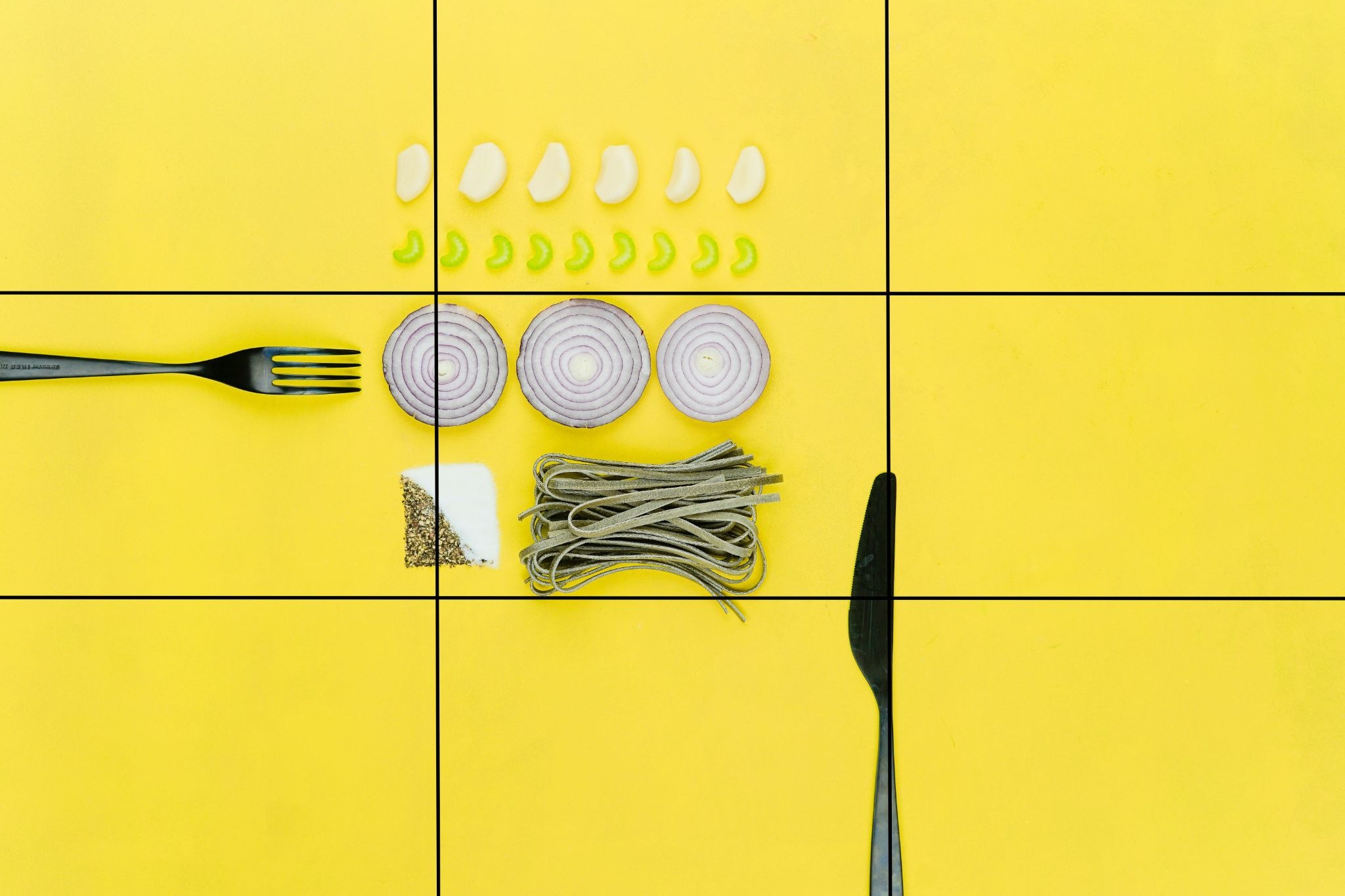 An overhead shot on a bright yellow background divided by a black grid shows neatly arranged ingredients including garlic cloves, celery slices, red onion rings, green fettuccine pasta, and a square of salt and pepper, flanked by a black fork and knife.
