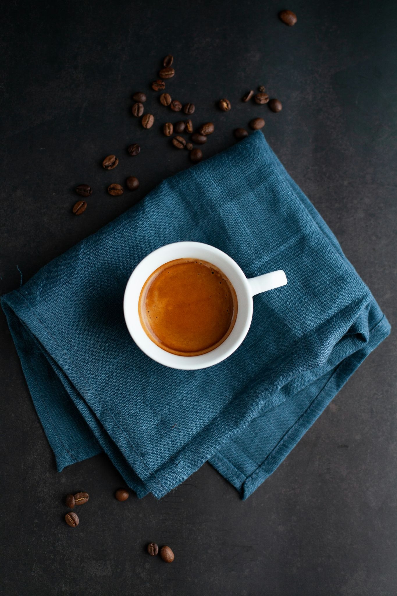 An overhead shot of a white cup filled with espresso, resting on a folded blue linen napkin surrounded by scattered coffee beans on a dark, textured surface.