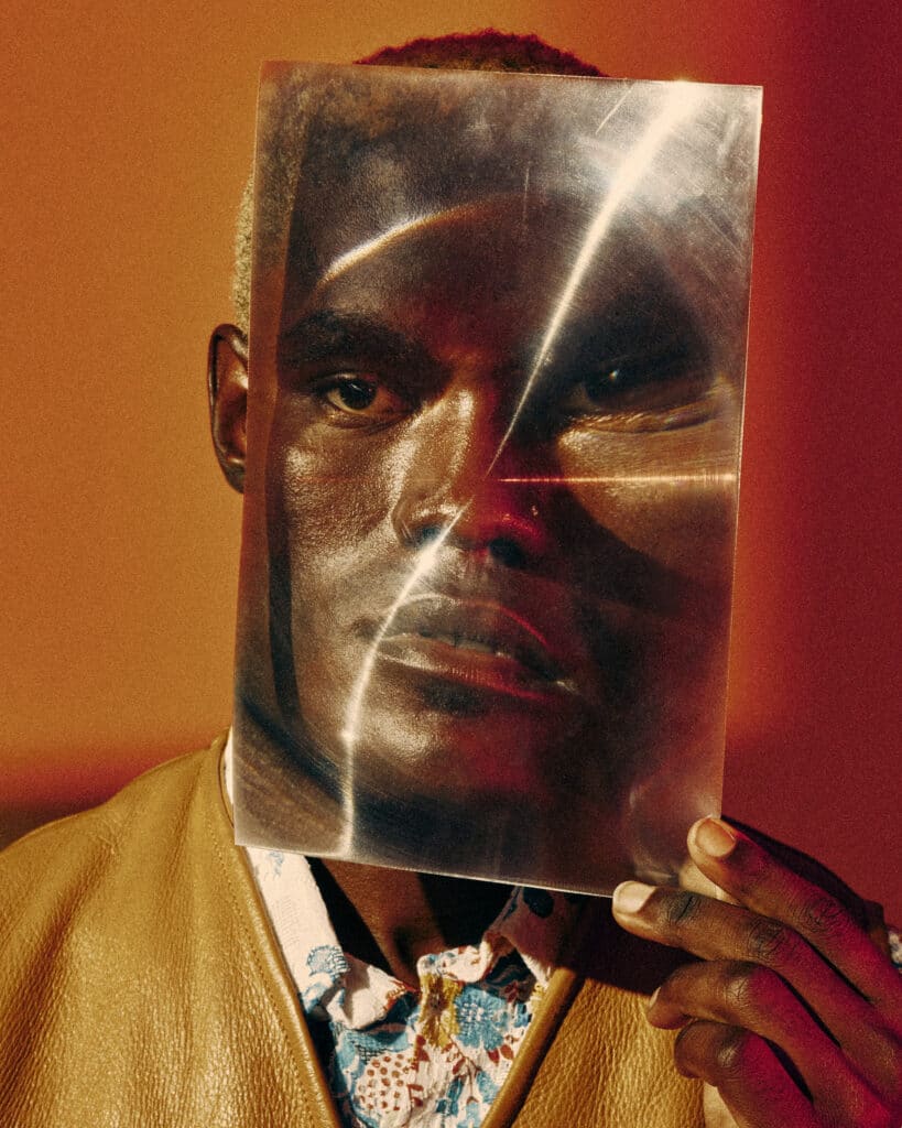 portrait of a black man wearing a mustard yellow leather jacket with a colorful patterned collared shirt underneath. he is holding a large rectangular clear filter in front of his face, causing his features to appear distorted