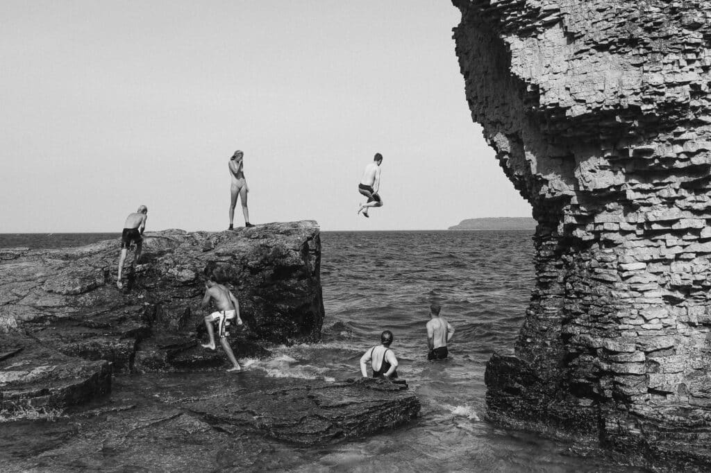 a black and white image of large rocky outcroppings surrounded by water, and people both swimming and waiting jumping off the rocks into the water below.