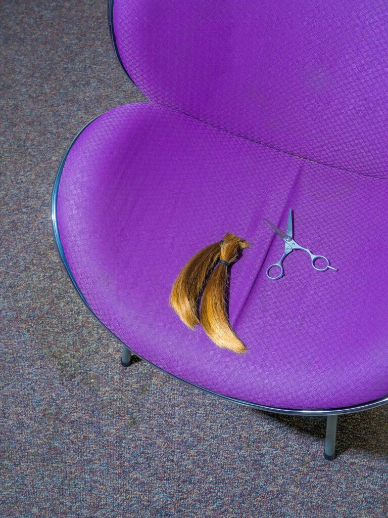 imagen desde arriba de una silla púrpura moderna sobre una alfombra gris con un par de tijeras pequeñas y dos secciones atadas de pelo, como los extremos de una coleta, descansando en el asiento