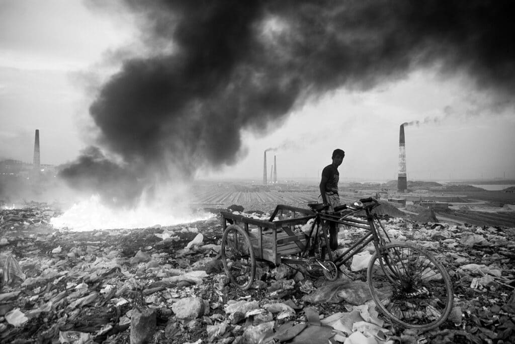 black and white image of an open space with trash all over the ground and several smokestacks in the distance. On the left, some of the trash is on fire, and a person stands to the right beside their makeshift bicycle carriage.