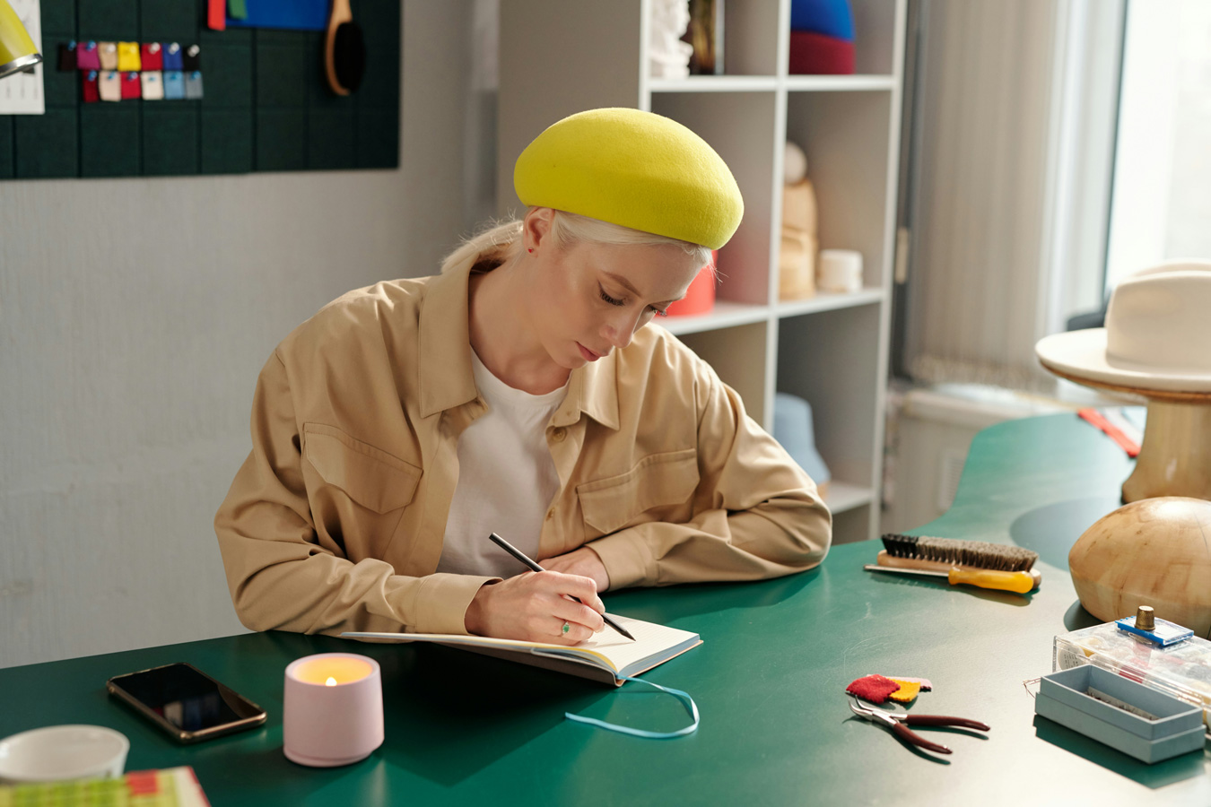 A woman in a yellow beret sits at a green table, writing in a notebook. Crafting tools and a lit candle are on the table. A shelf with hat-making supplies is in the background.