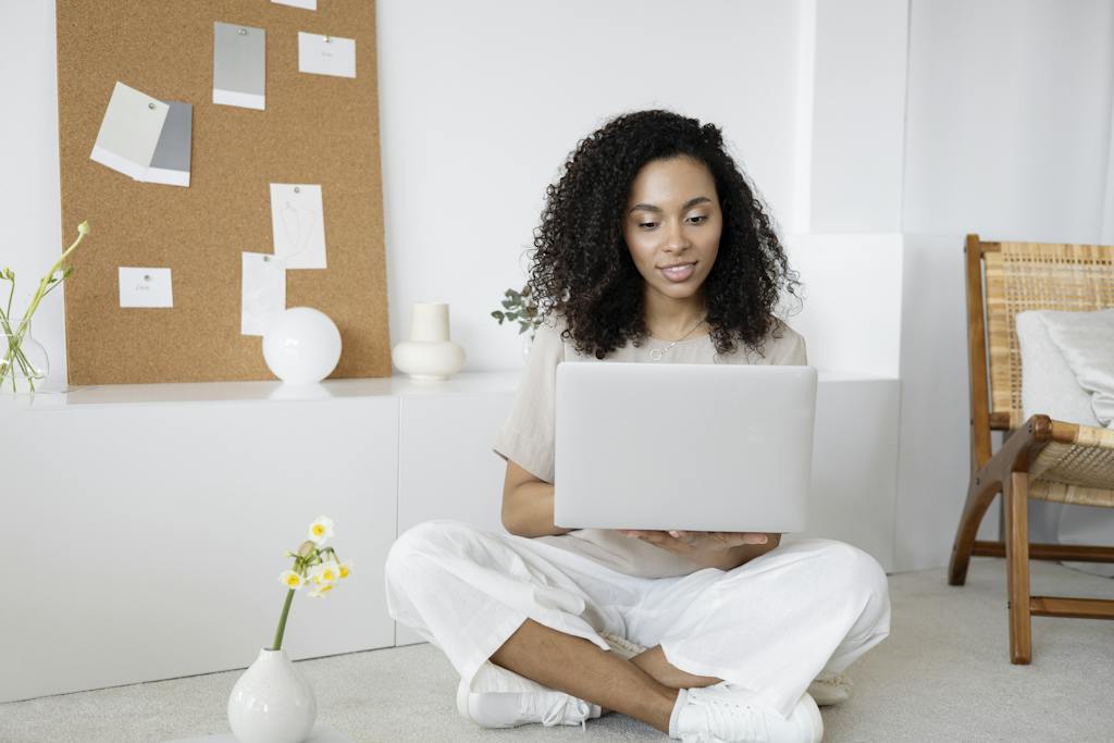 Mulher de blusa bege e calça branca sentada no chão usando um Macbook