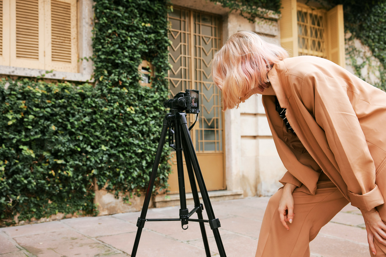 woman with pink hair wearing a beige suit looking at the screen on a digital camera on a tripod